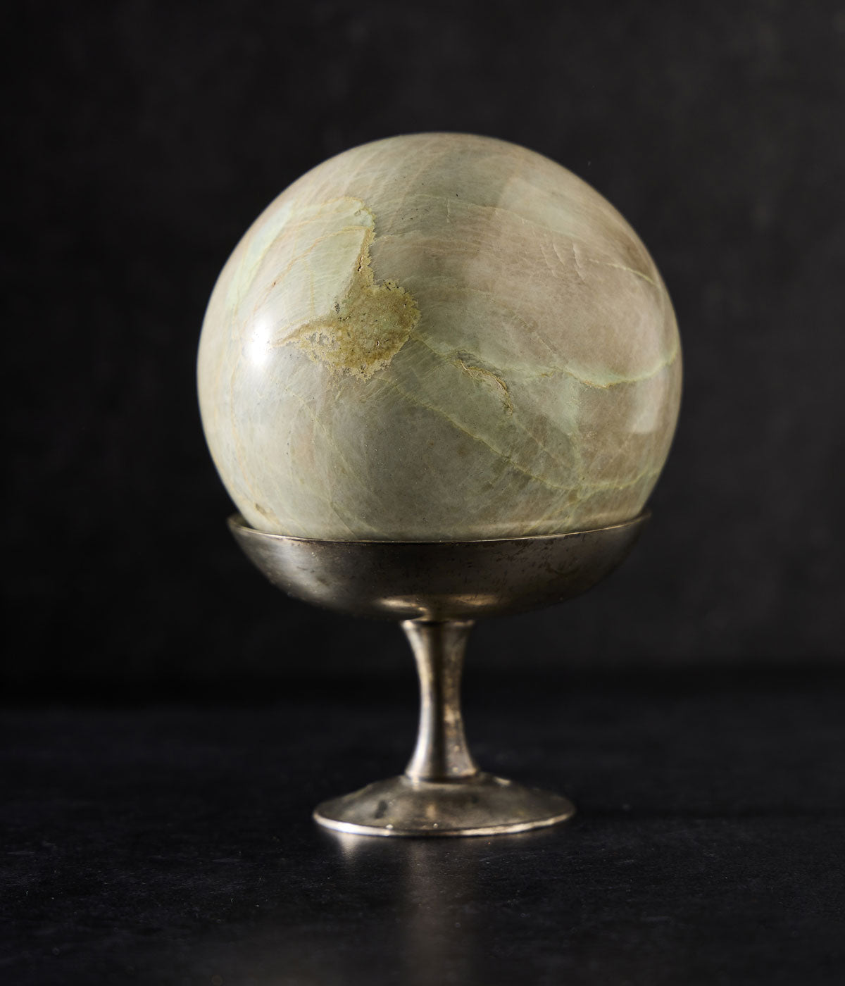 A polished Garnierite Green Moonstone Sphere with a metallic stand, displayed against a dark background.