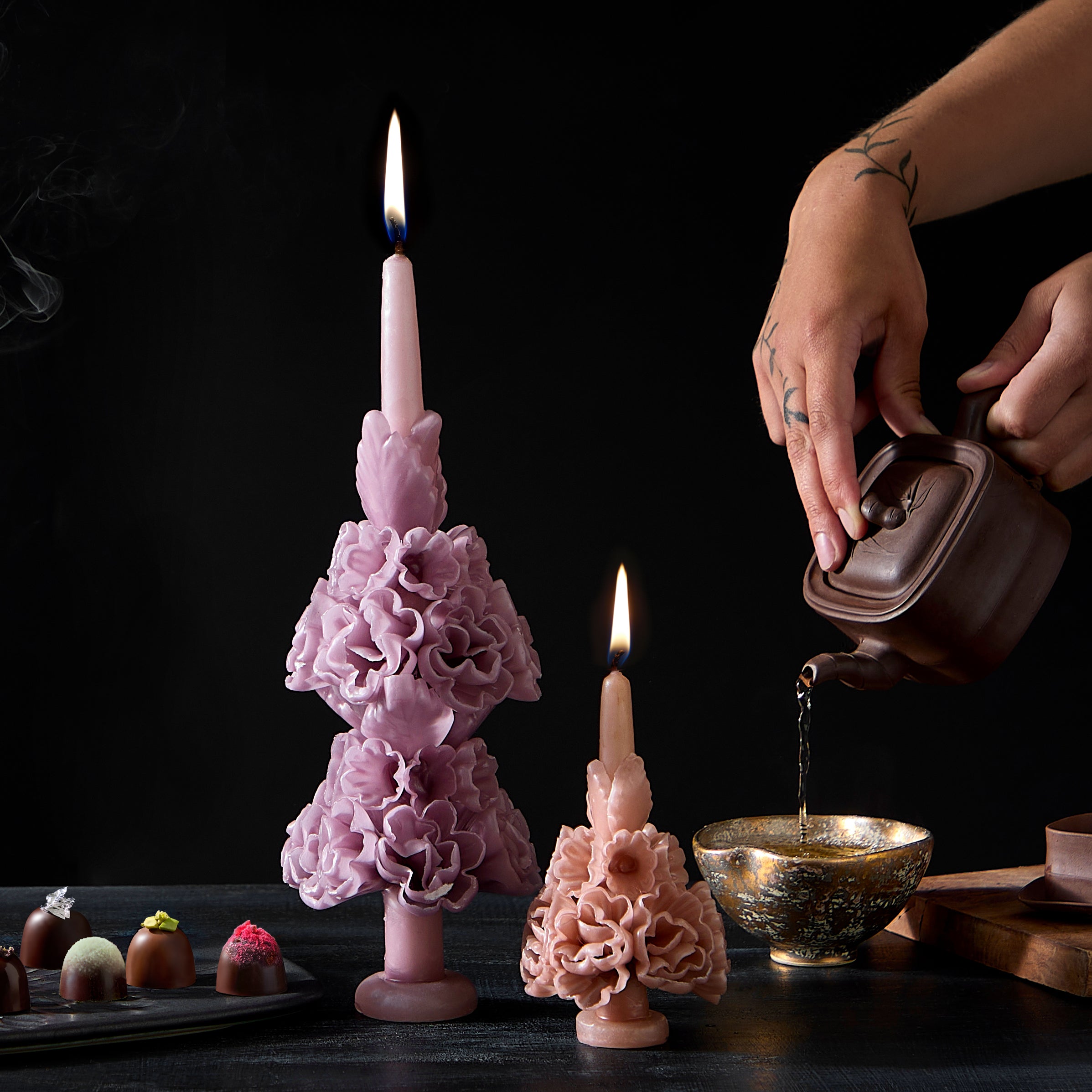 Lavender and Pink floral candles next to a pot of tea being poured. Chocolate Truffles on the table.