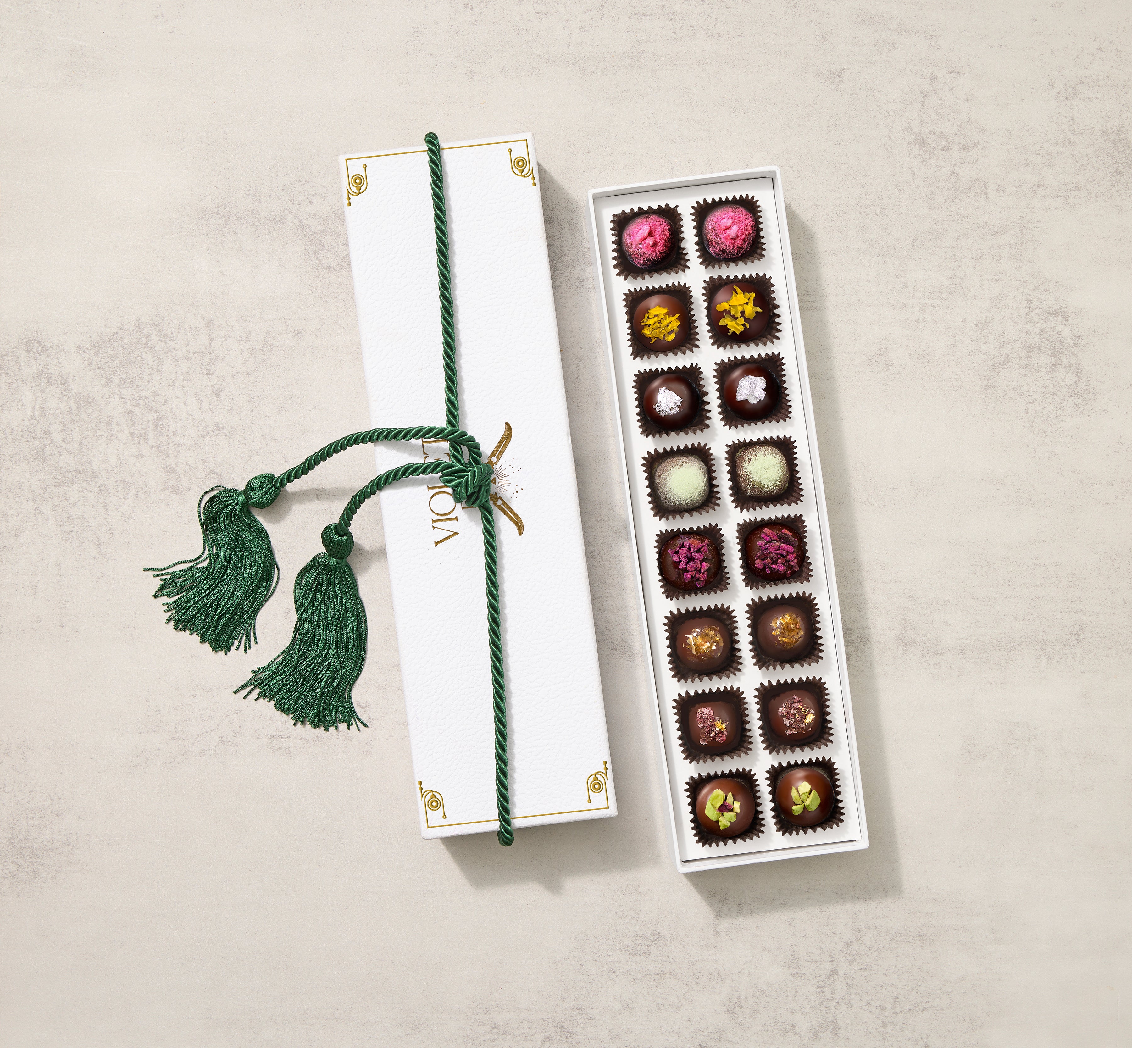 A gift box of assorted milk chocolate and dark chocolate truffles from Violet Flame Chocolate, featuring a variety of colors and designs, tied with a decorative green ribbon.