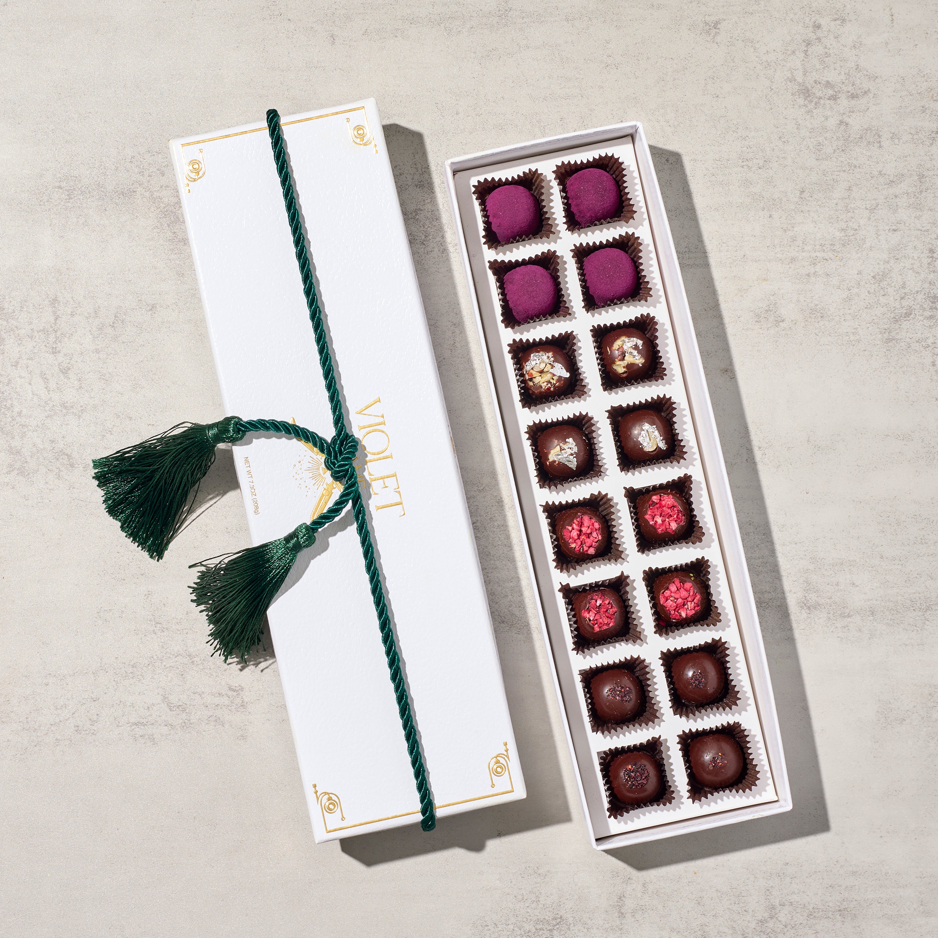 A gift box of dark chocolate truffles topped with various artisanal ingredients presented in a beautifully packaged box with a green decorative tassel.