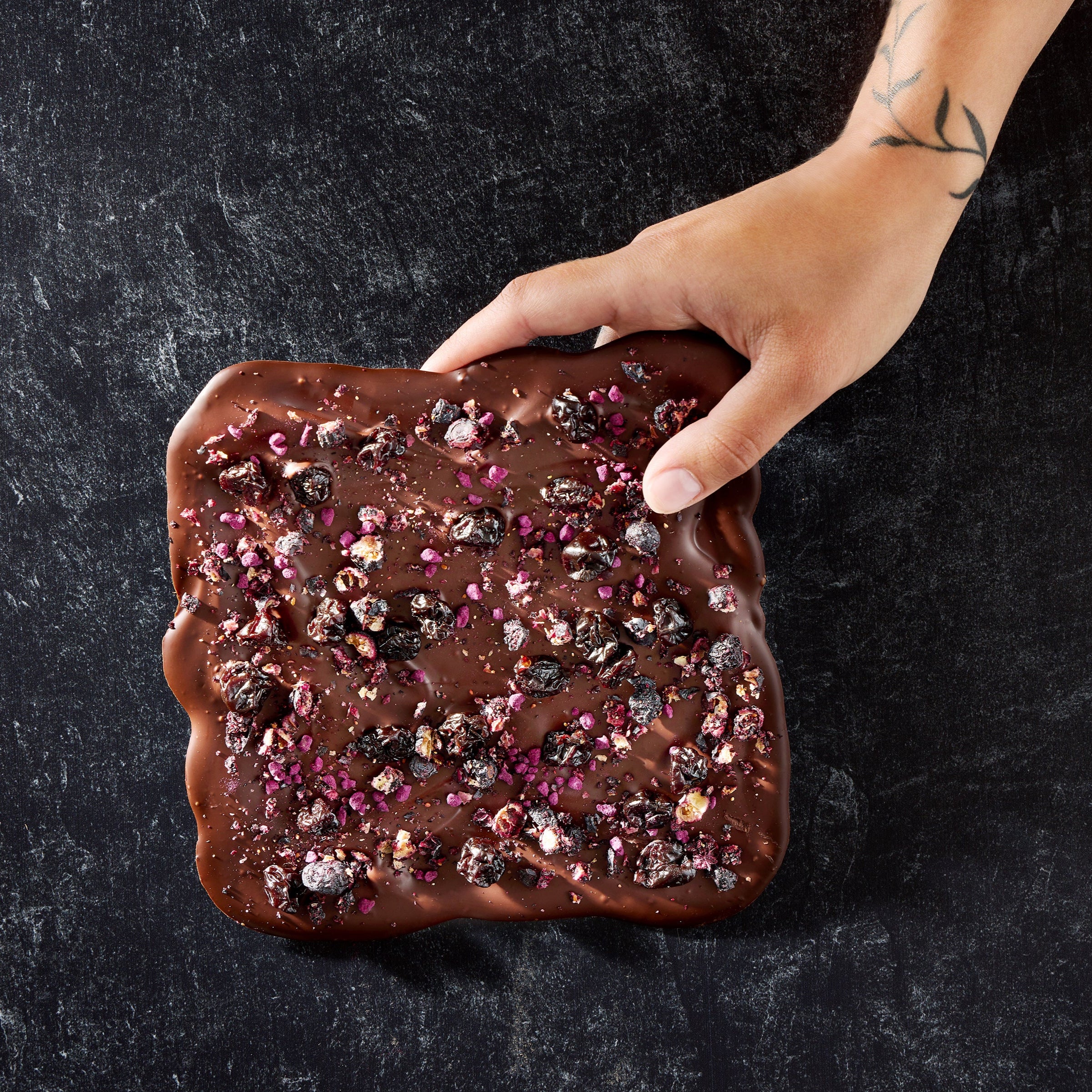 A close-up image of a dark chocolate board with a layer of dried fruit pieces and a sprinkle of Maldon sea salt on top, held by a person.