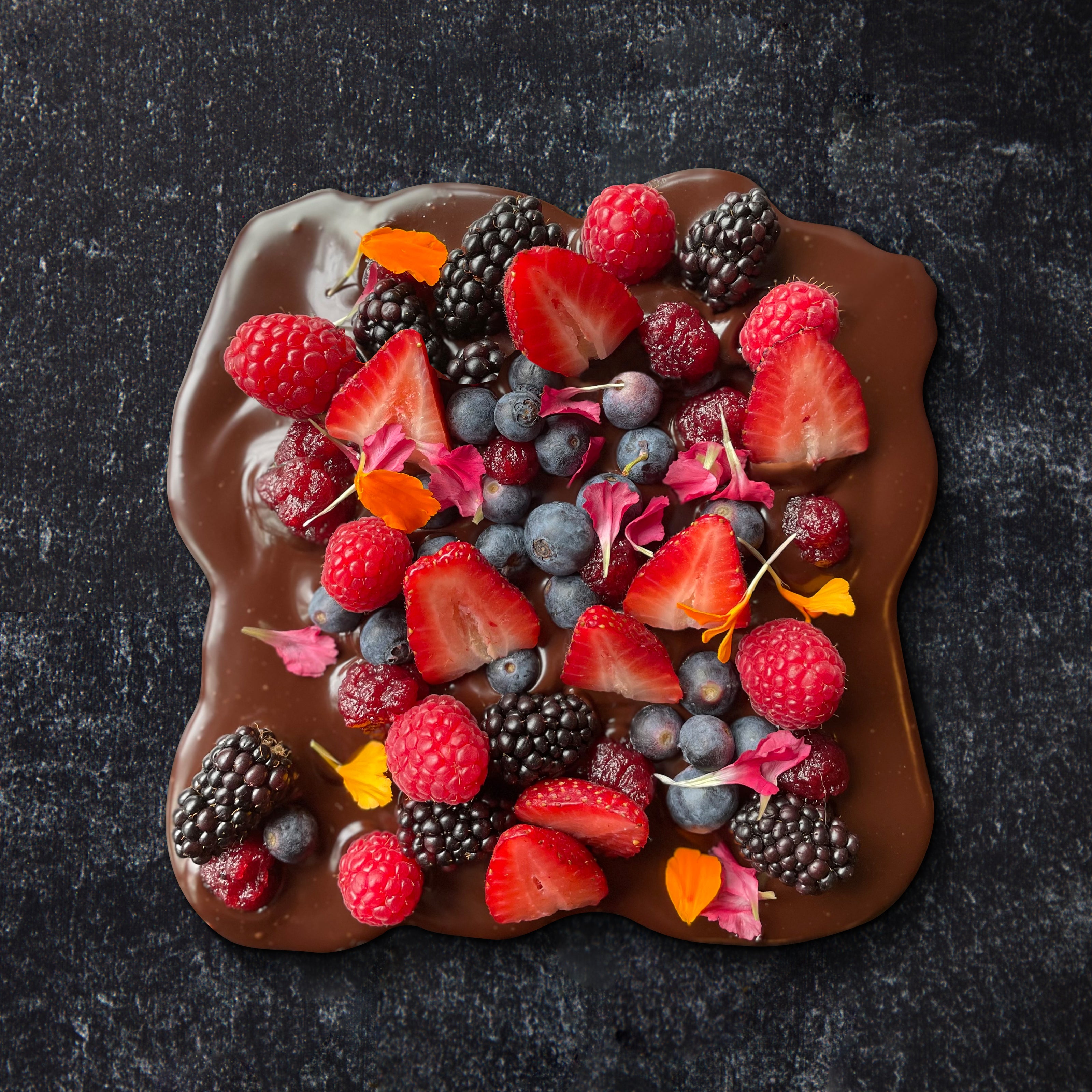 A chocolate ganache board topped with a variety of fresh strawberries, blueberries, raspberries, and edible flowers, served on a dark surface.