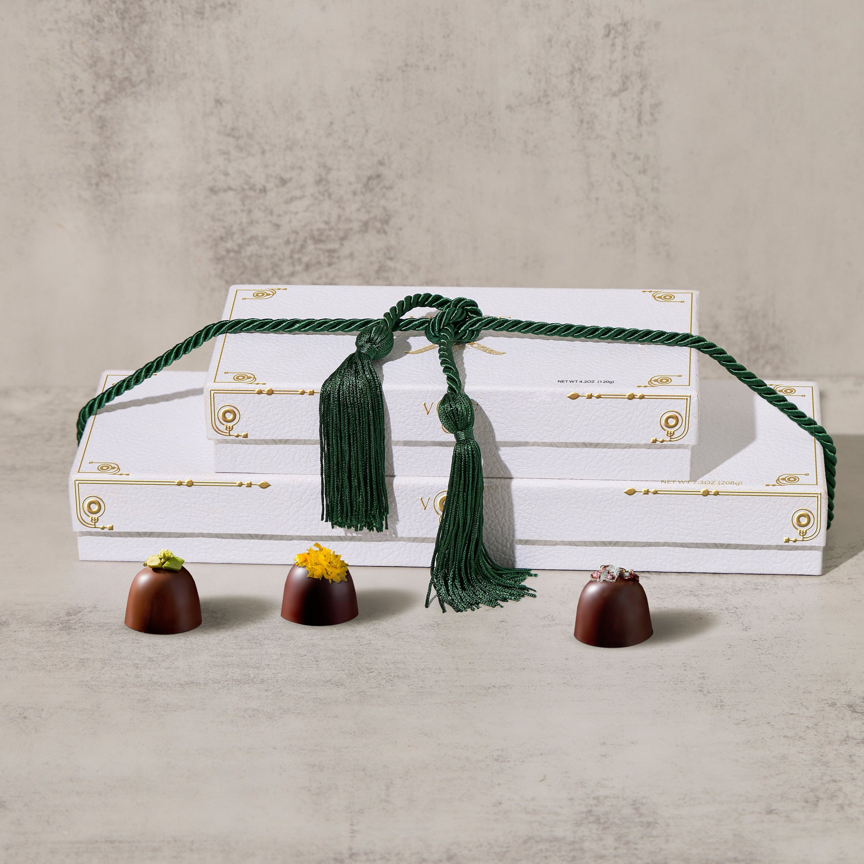 A white box gift tower of assorted chocolates with a green decorative tassel, displayed with a few chocolates in front.
