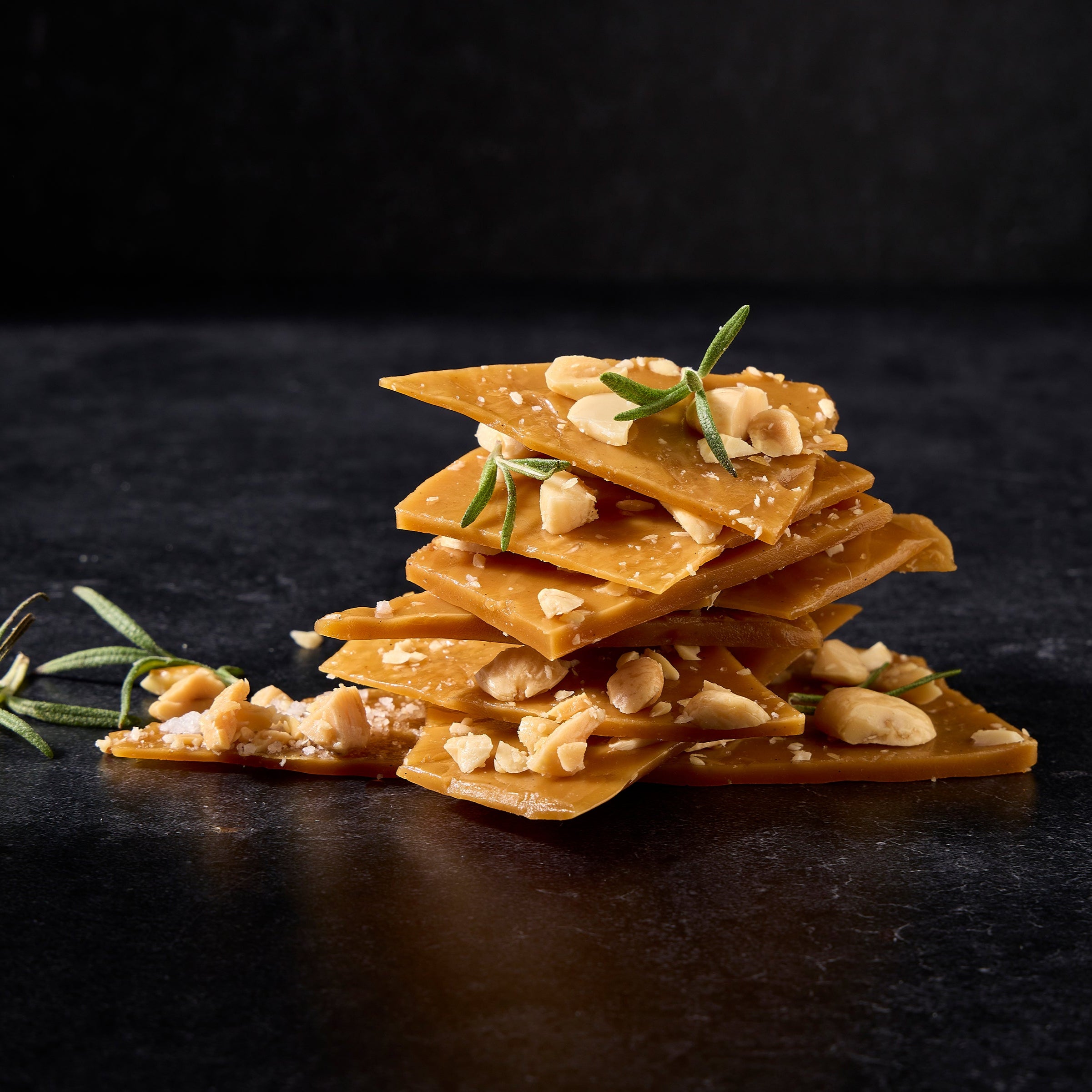 Stack of toffee pieces with visible toasted almonds and a sprig of rosemary, on a dark background.