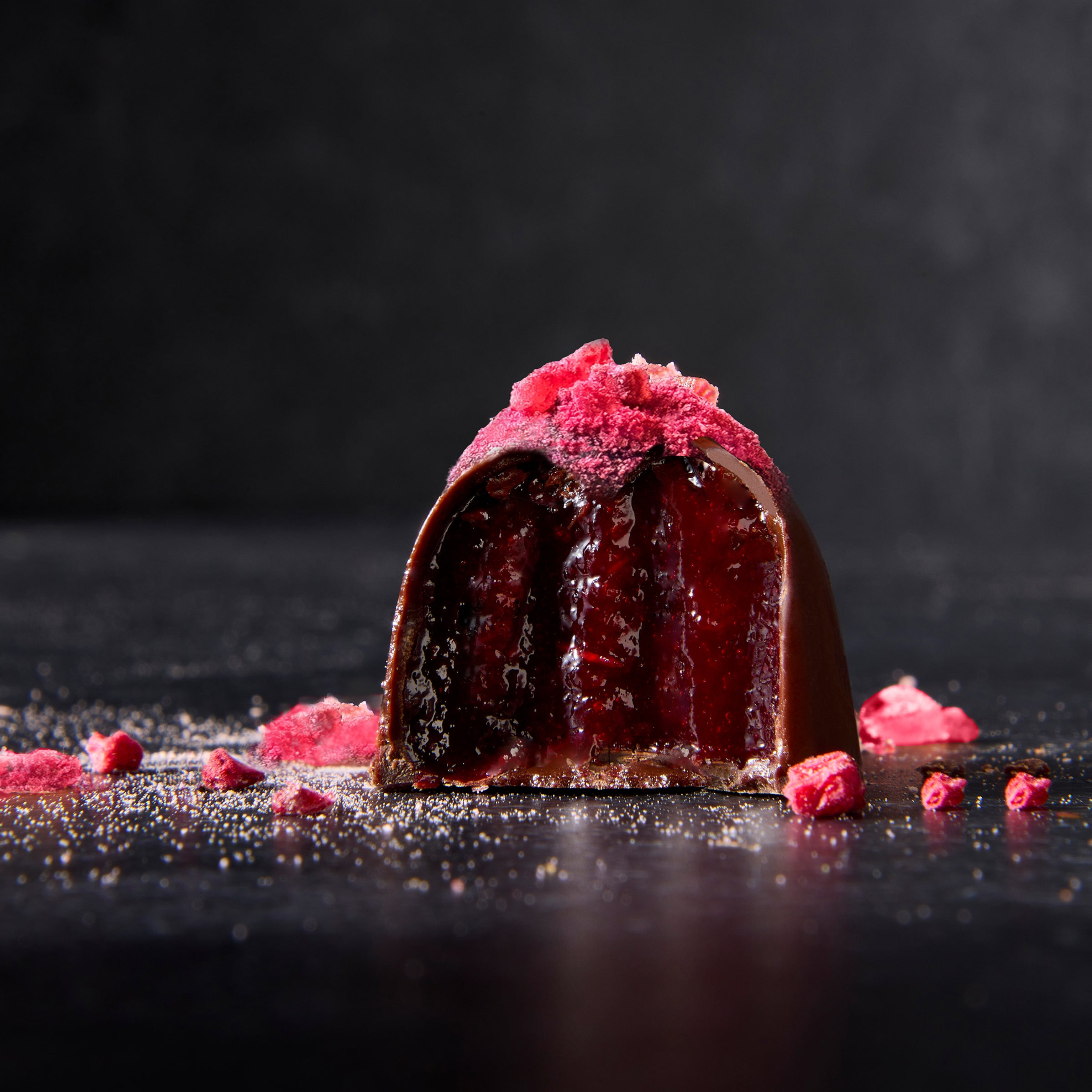 A chocolate truffle with a pink, glossy exterior and bits of raspberries and spices scattered around on a dark background.