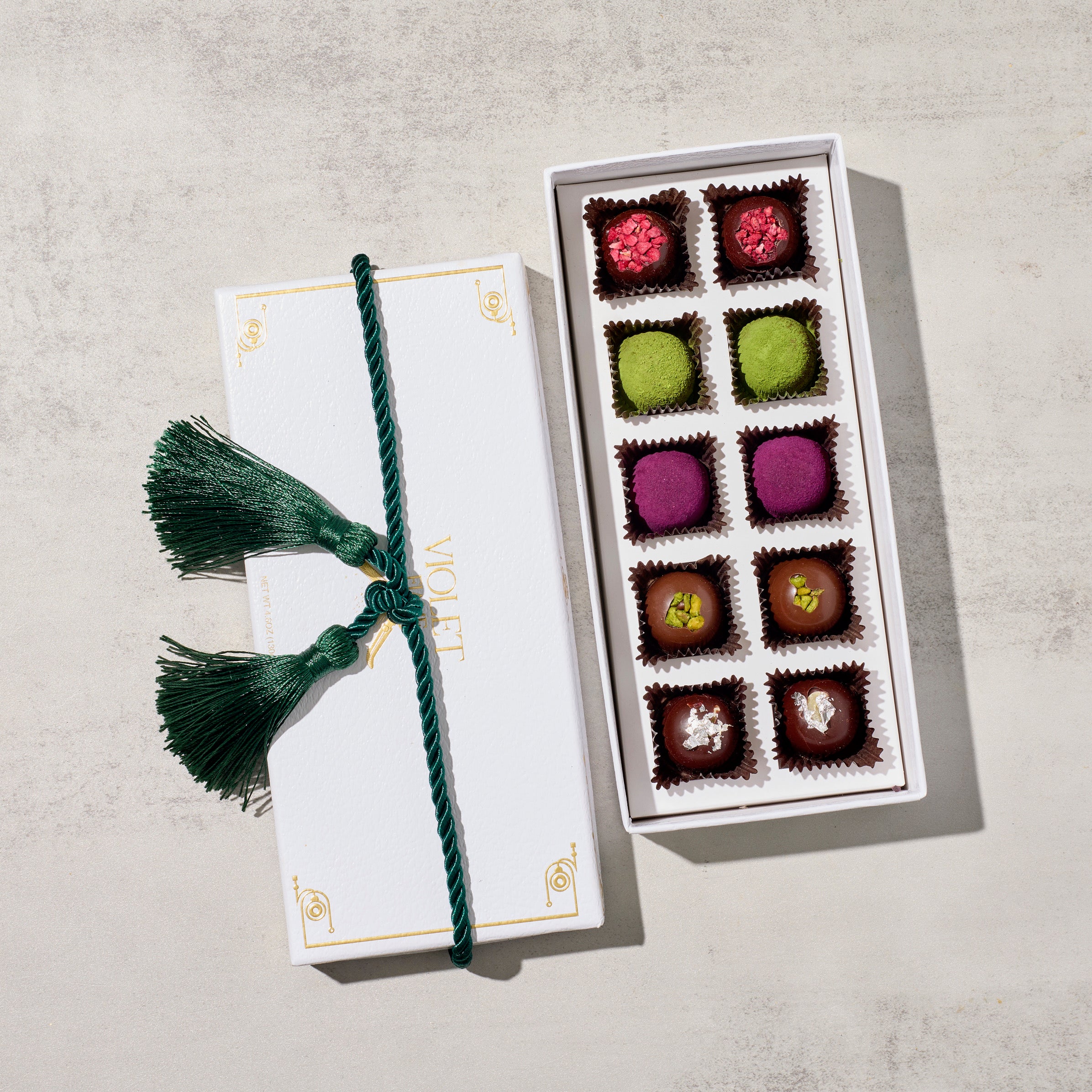 A gift box of assorted chocolates truffles, presented in a beautifully packaged box tied with a green tassel. The chocolates are visible through the open lid, displaying a variety of colors and textures. 